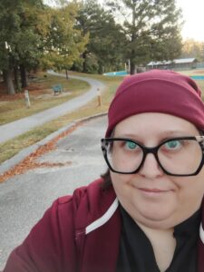 woman with burgundy beanie and hoodie and black cat eye glasses. A path is behind her. 